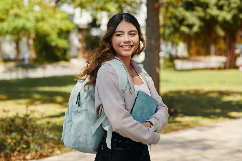 A young student smiling outdoors, holding a notebook and wearing a backpack